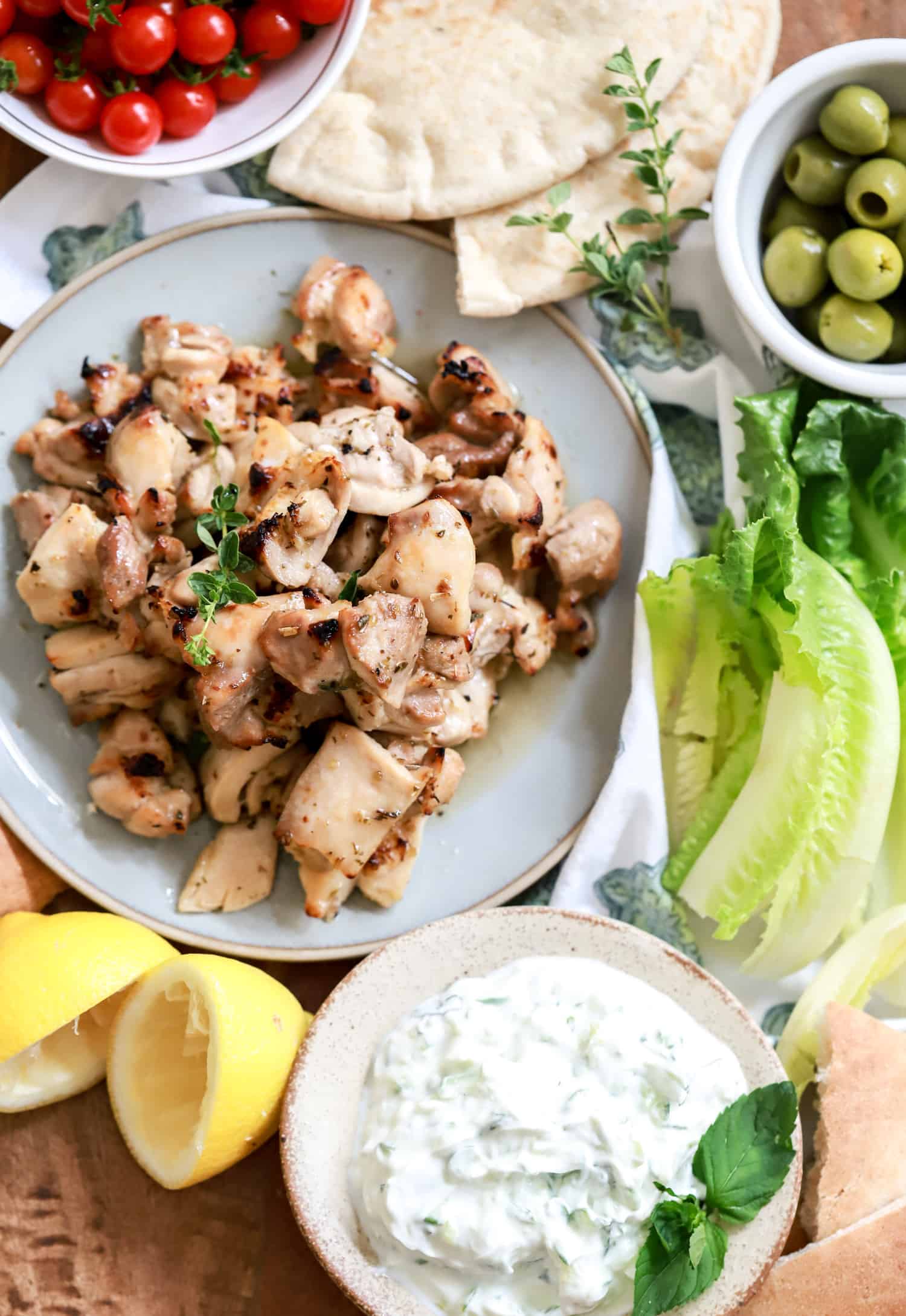crowded board with plate of chicken, lemon halves, tzatziki, pita, and lettuce leaves.