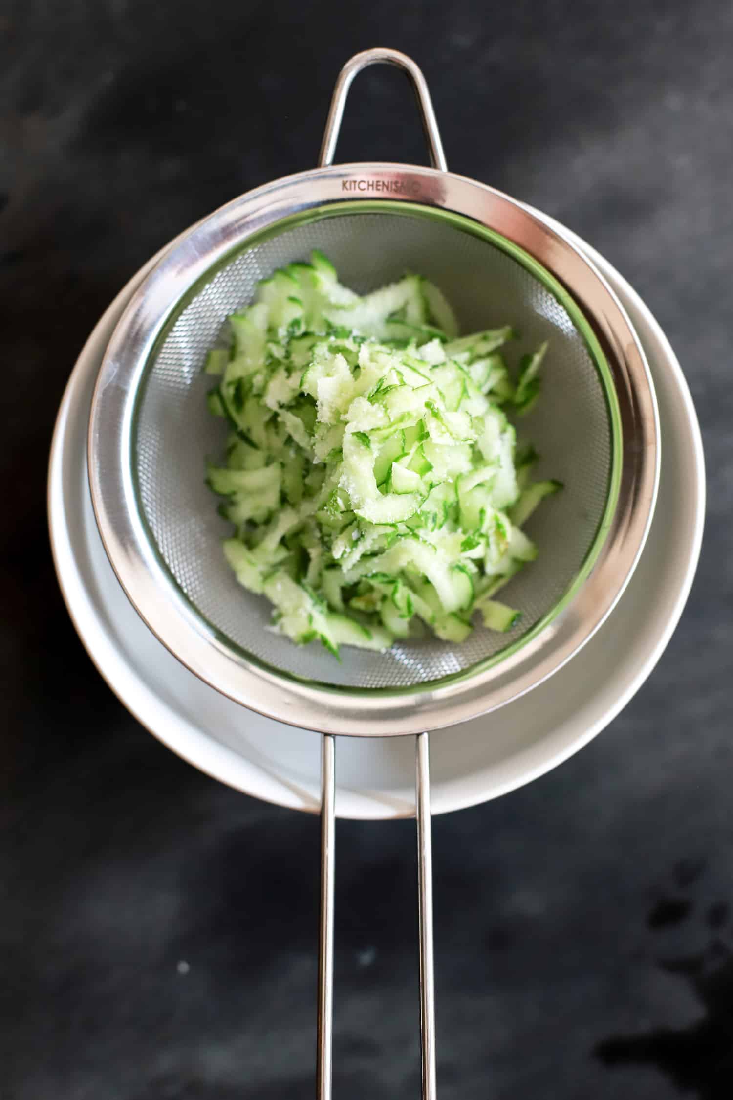 grated cucumber in fine mesh strainer over white bowl on black background