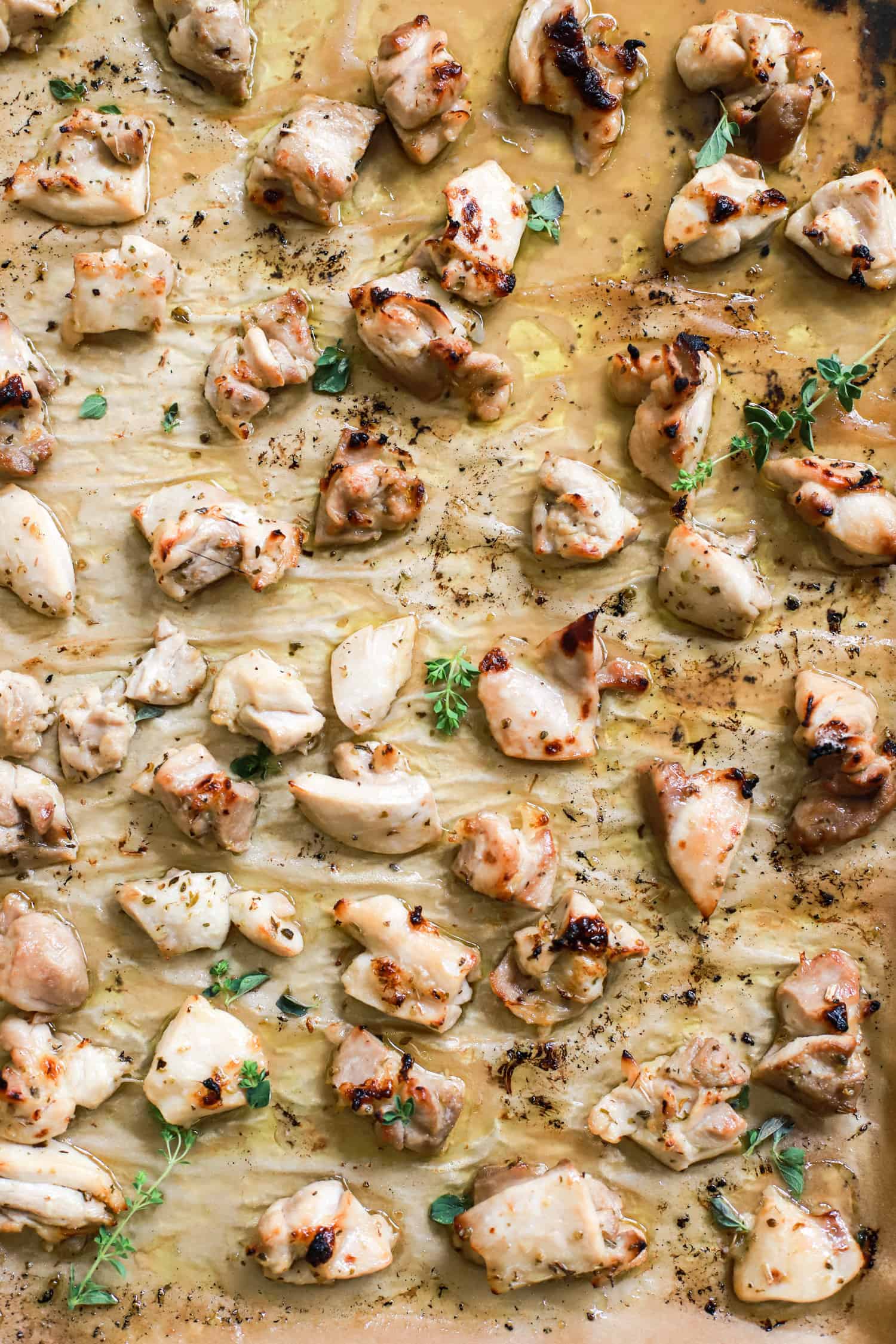 close up tray of broiled chicken bites with oregano leaves on brown parchment paper.