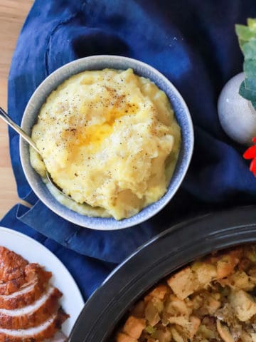 bowl of mashed potatoes with butter swirl on blue napkin surrounded by Thanksgiving foods.