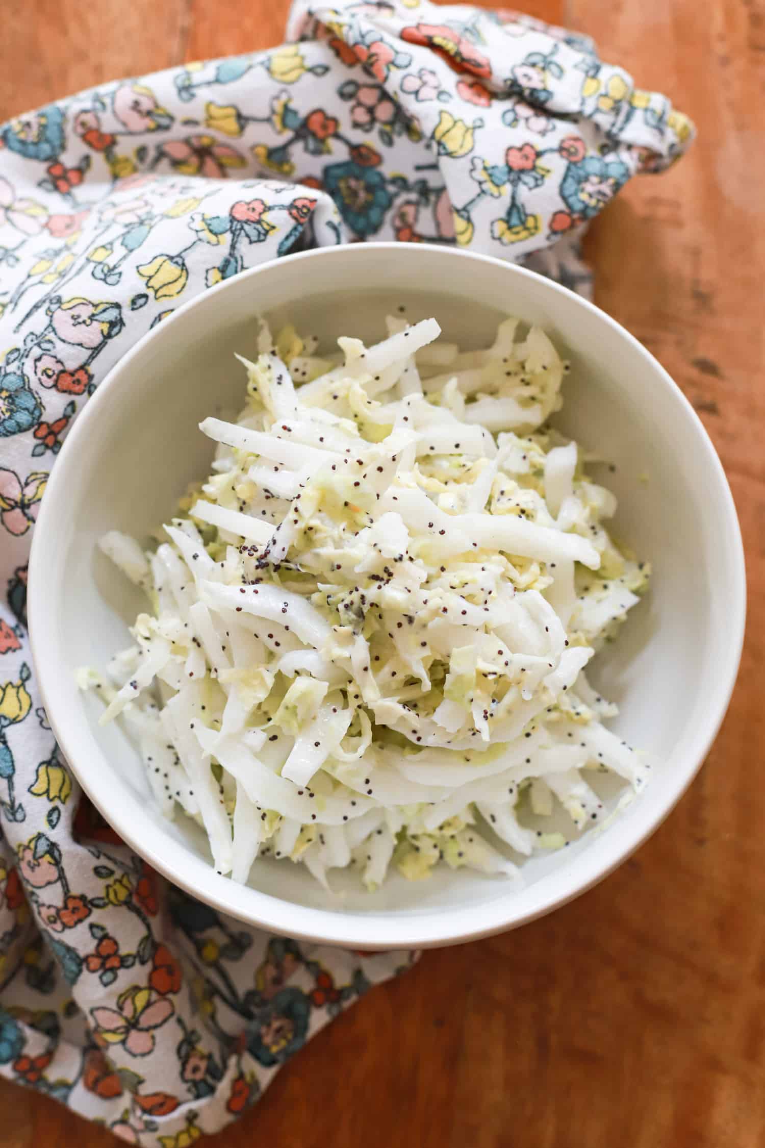 white bowl of poppyseed slaw with Greek yogurt on wooden board with floral napkin.