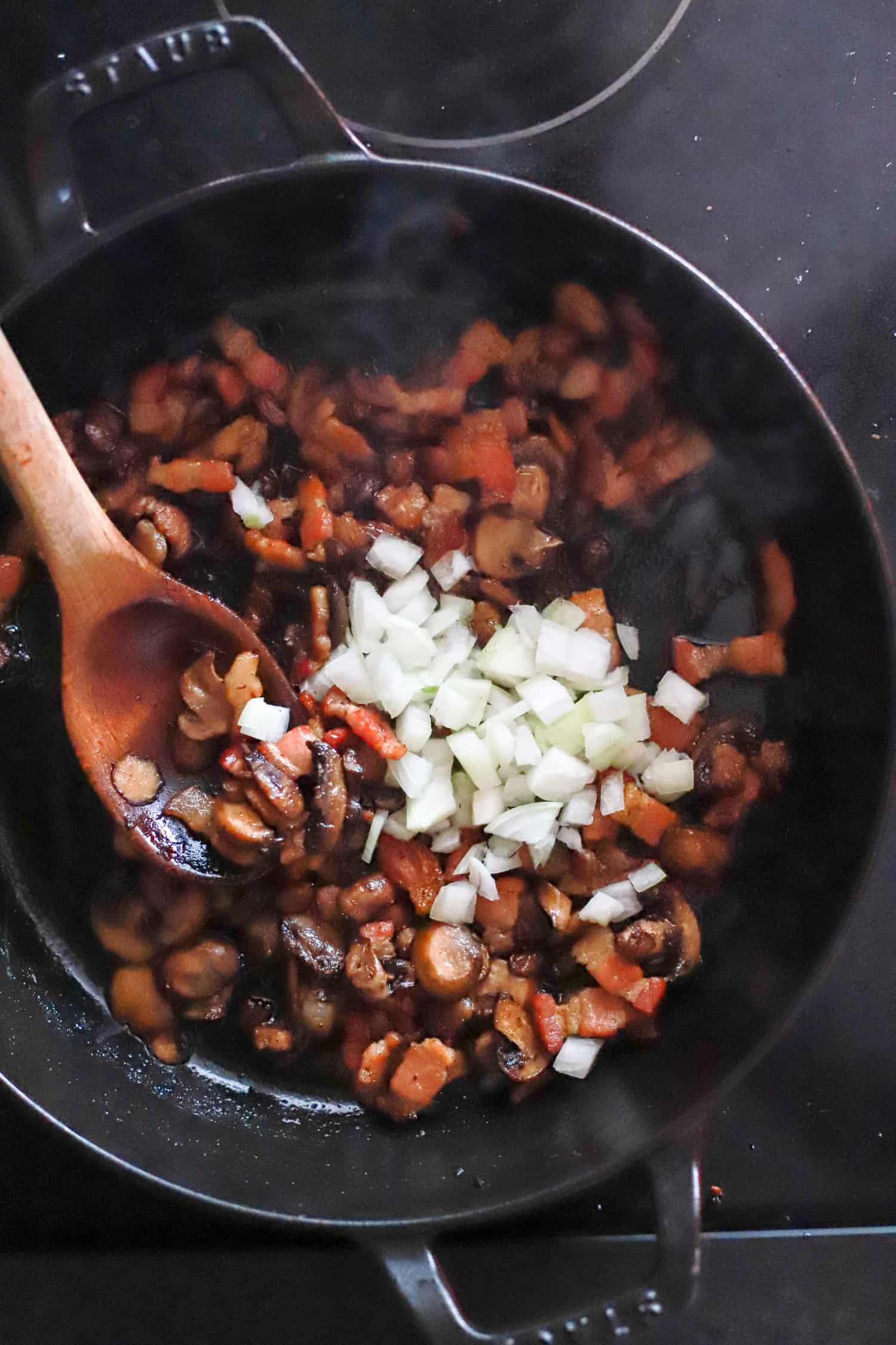 mushrooms, onions, and bacon crisping in a skillet with wooden spoon.