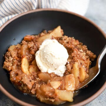 Black bowl of apple crisp (no oats) topped with vanilla ice cream.