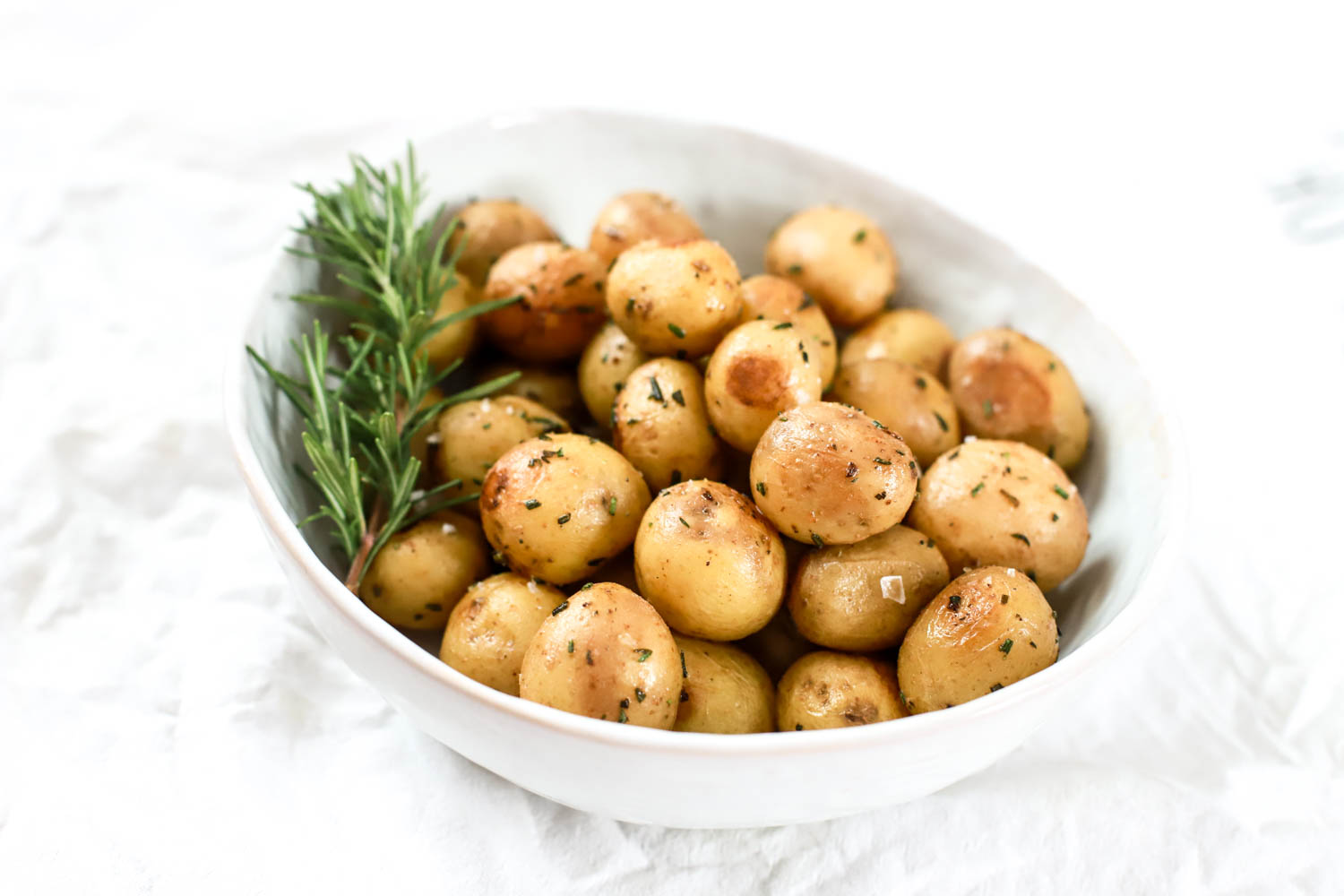 Bright white bowl of tiny potatoes and rosemary on a white cloth.