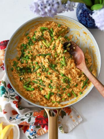 Skillet with spring veggie risotto surrounded by floral napkin and purple flowers.