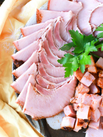 Sliced ham on a platter with herbs and a yellow napkin.