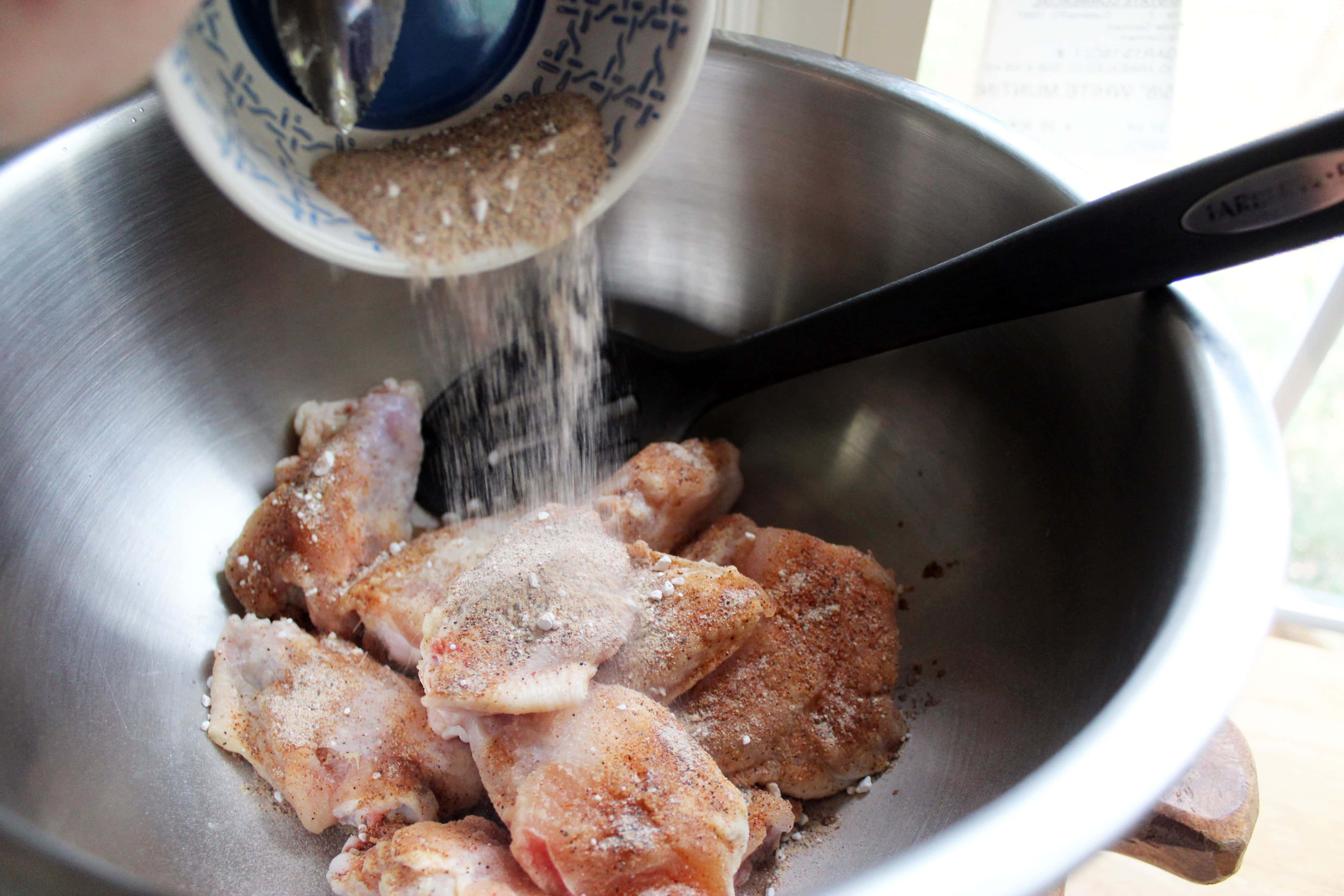silver bowl with chicken wings and dry rub spices being poured over wing pieces.