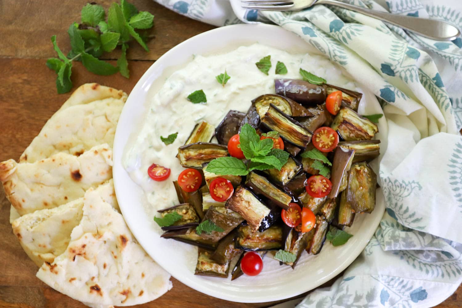 plate of yogurt with roasted eggplant and yogurt plus naan on board with decorative napkin