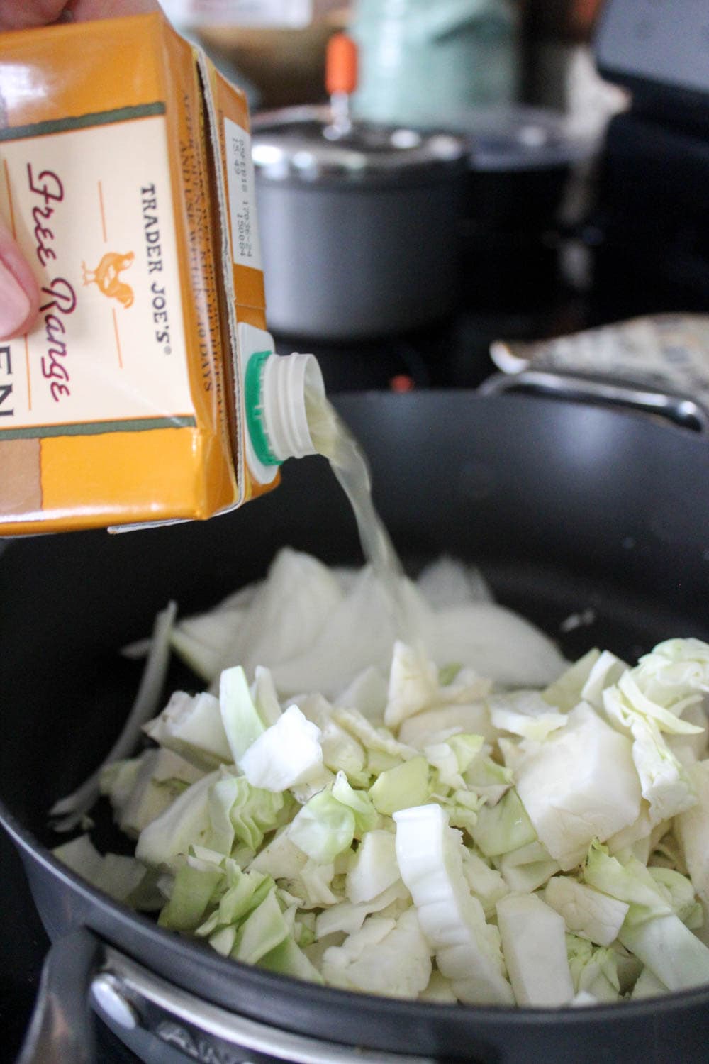 Box of chicken stock being poured into a pan with chopped green cabbage.