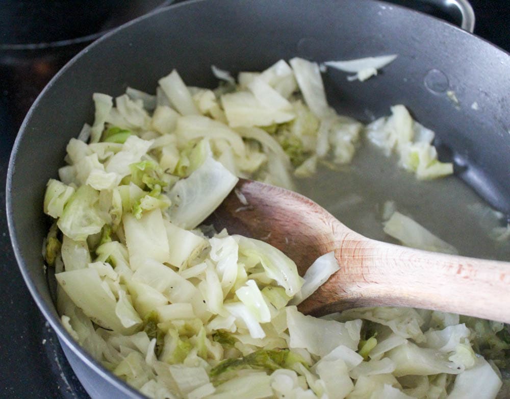 Gray skillet with wooden spoon stirring stewed cabbage.