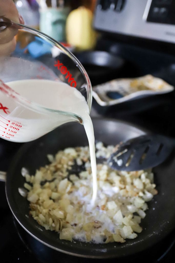 glass measuring cup pouring milk into skillet with onions and roux.