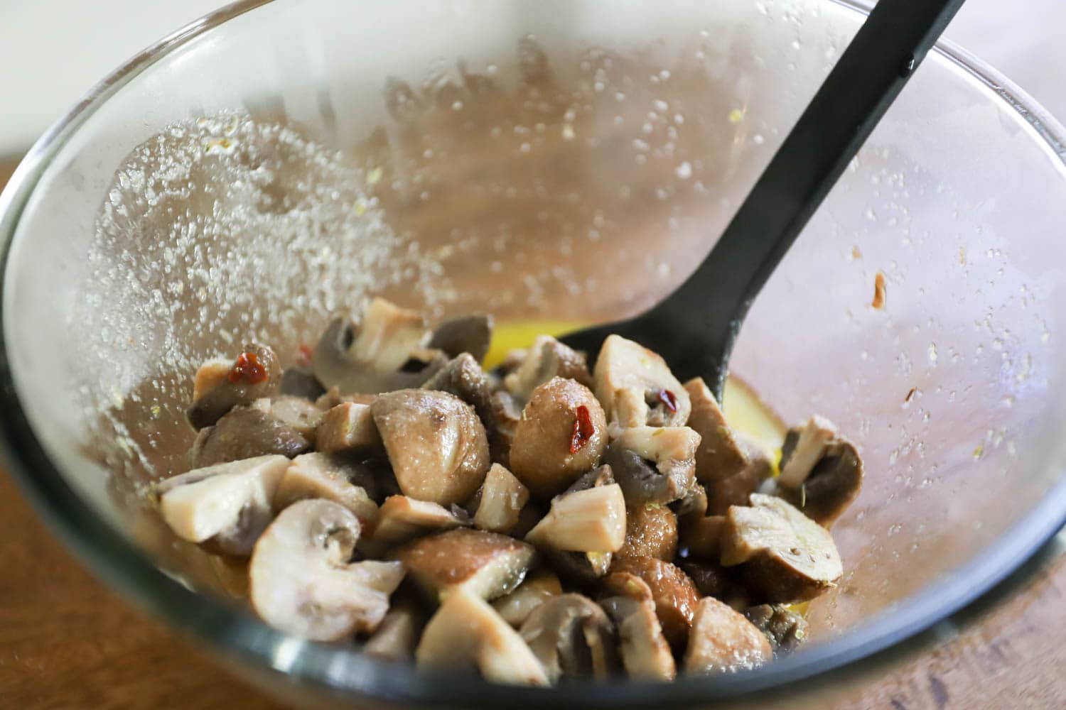 mushrooms stirred into marinade with black plastic spoon in glass bowl.