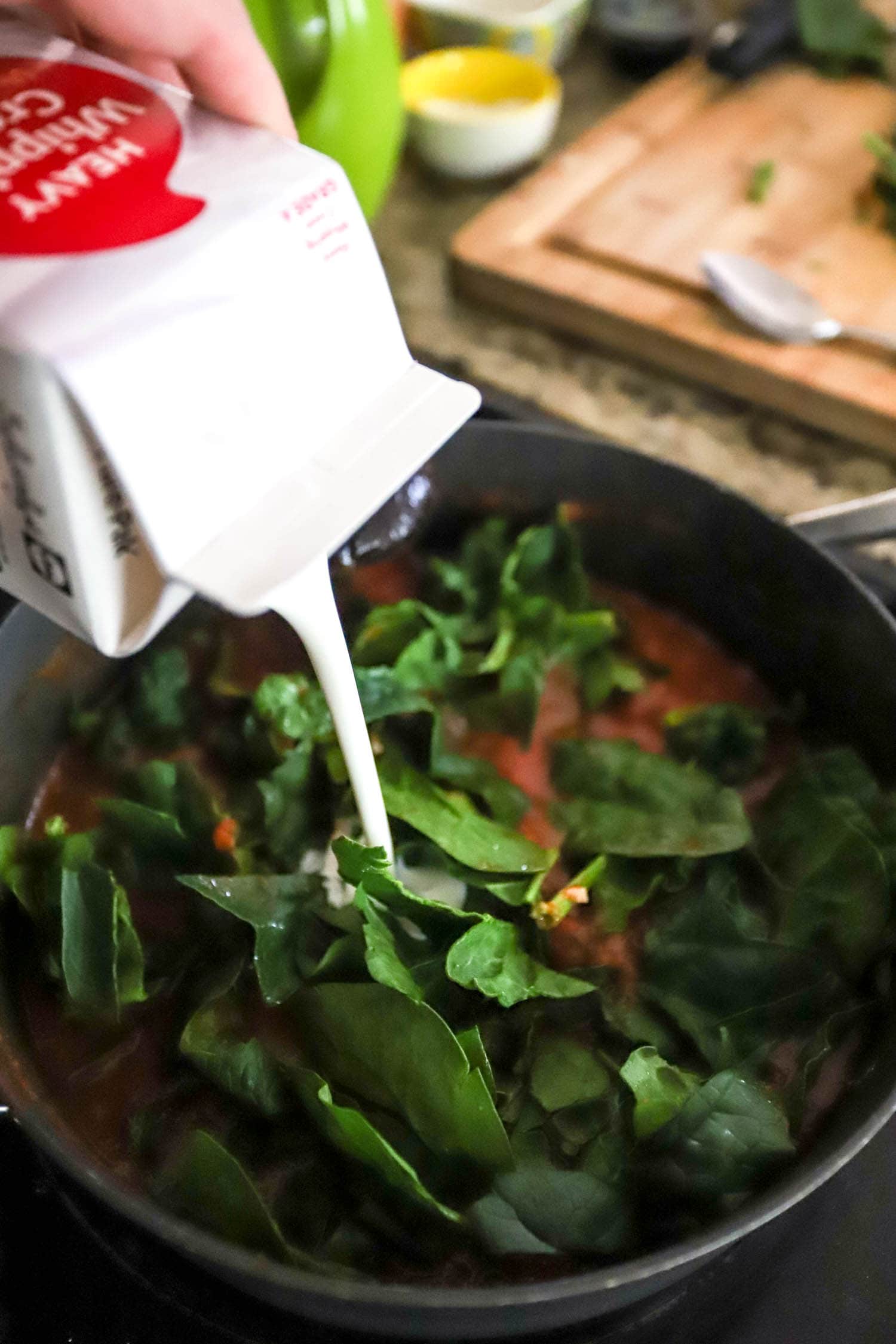 hand pouring heavy cream into skillet with spinach and tomato cream sauce.