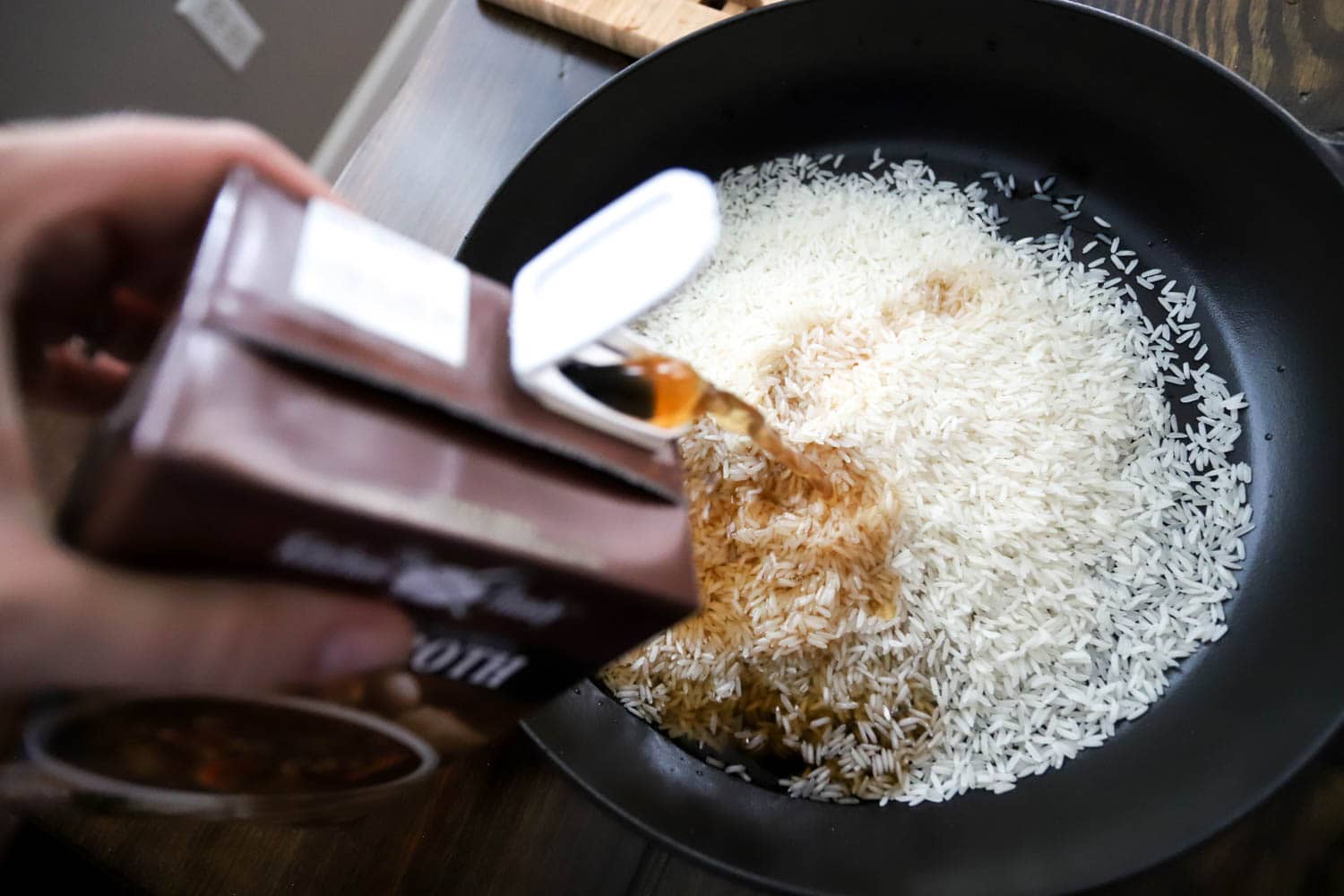 hand pouring beef broth over raw rice.