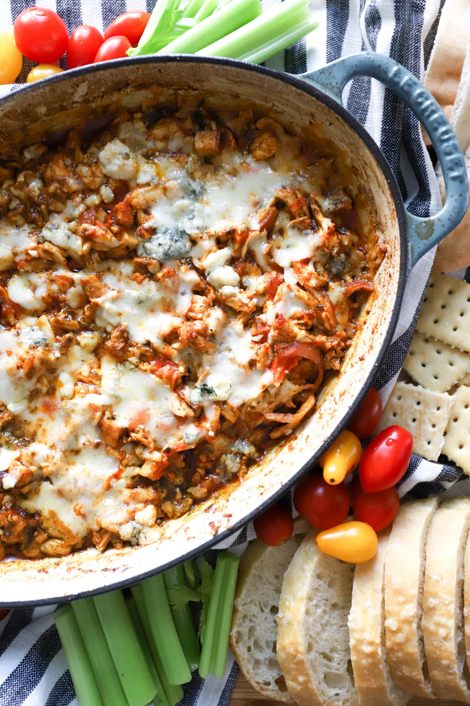 Hot buffalo chicken dip with melted cheese on top in blue skillet surrounded by veggies.