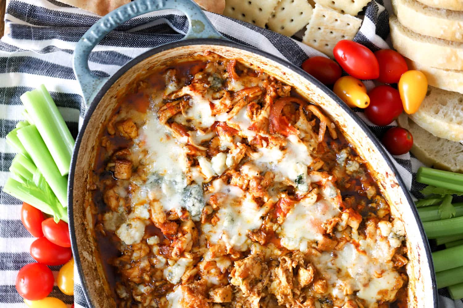 blue baking dish with buffalo chicken dip surrounded by fresh veggies, crackers, and bread.