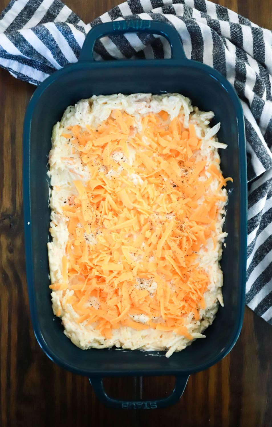 unbaked hashbrown casserole topped with cheese in dark green baking dish with striped napkin.