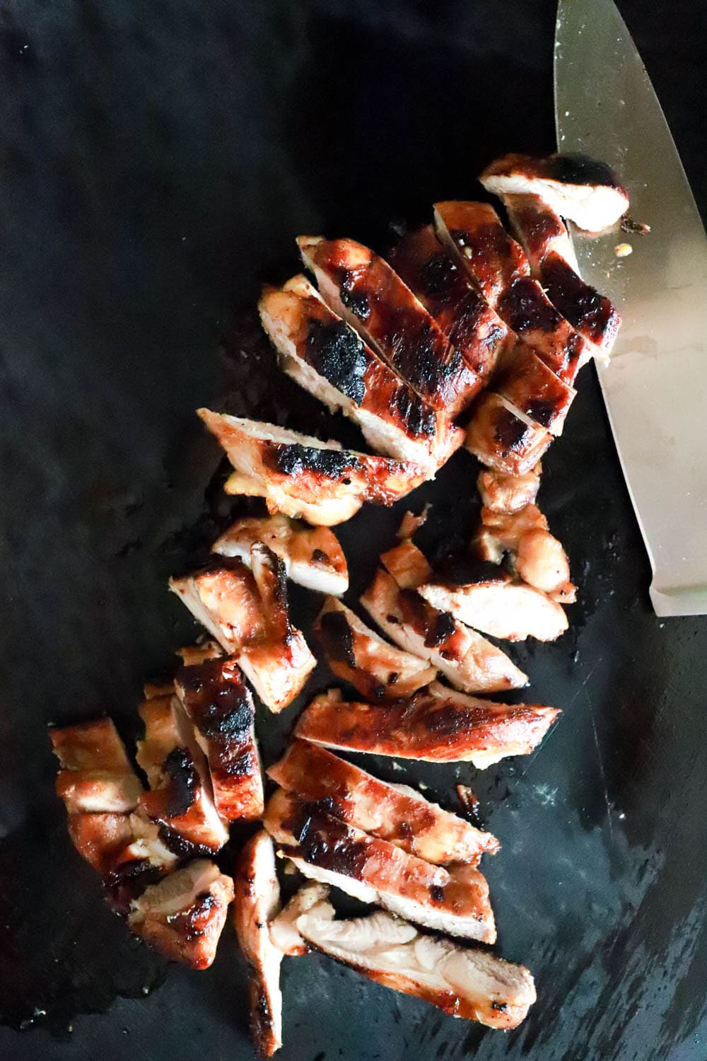 Sliced grilled chicken with a large chef's knife on a black cutting board.