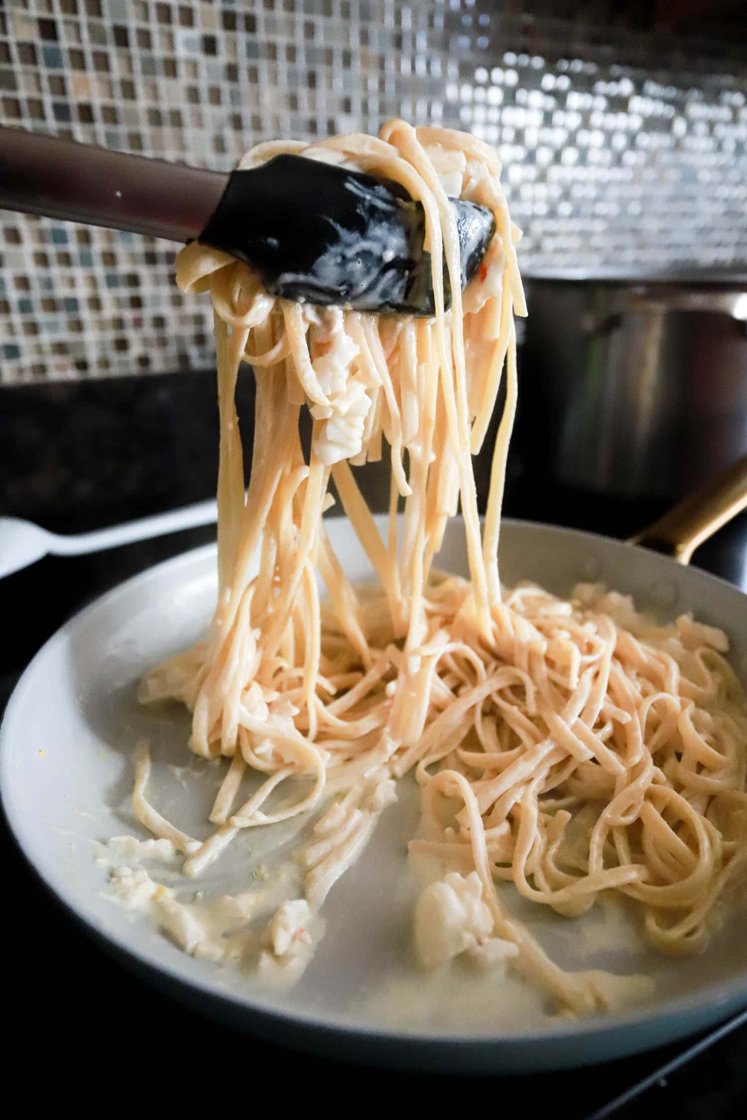 Tongs tossing creamy lobster pasta in large skillet.