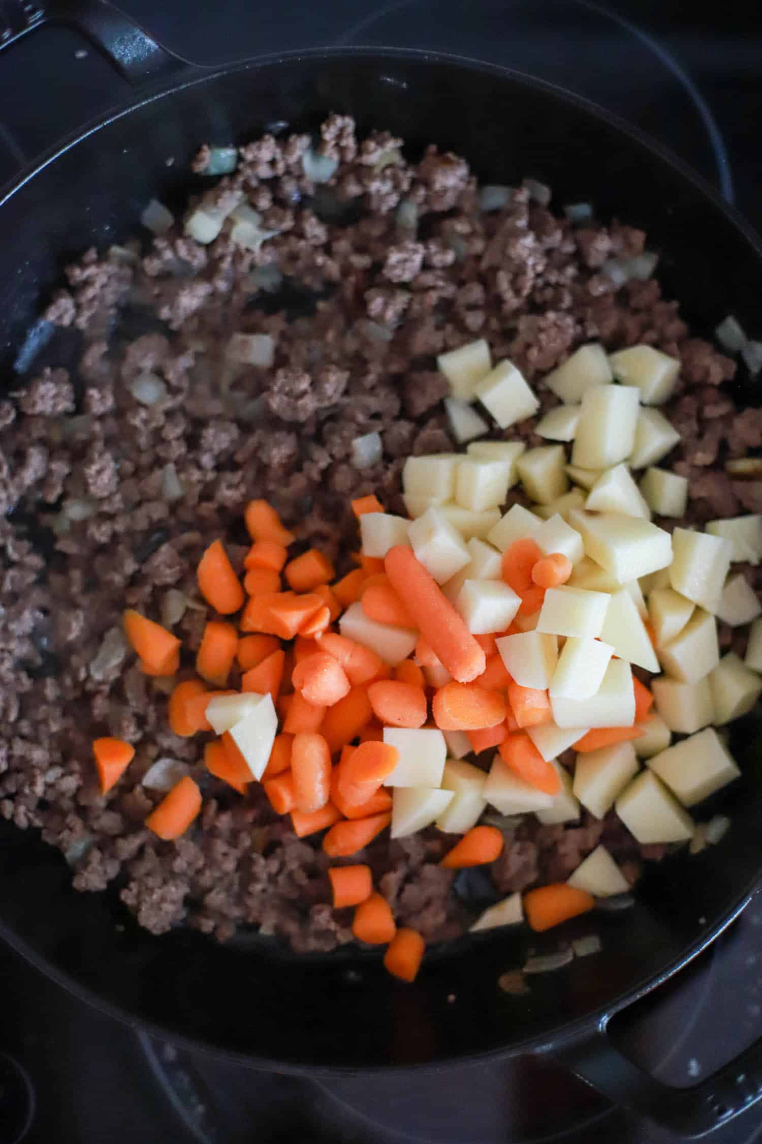 Carrots and potatoes on top of ground beef in a large black skillet.