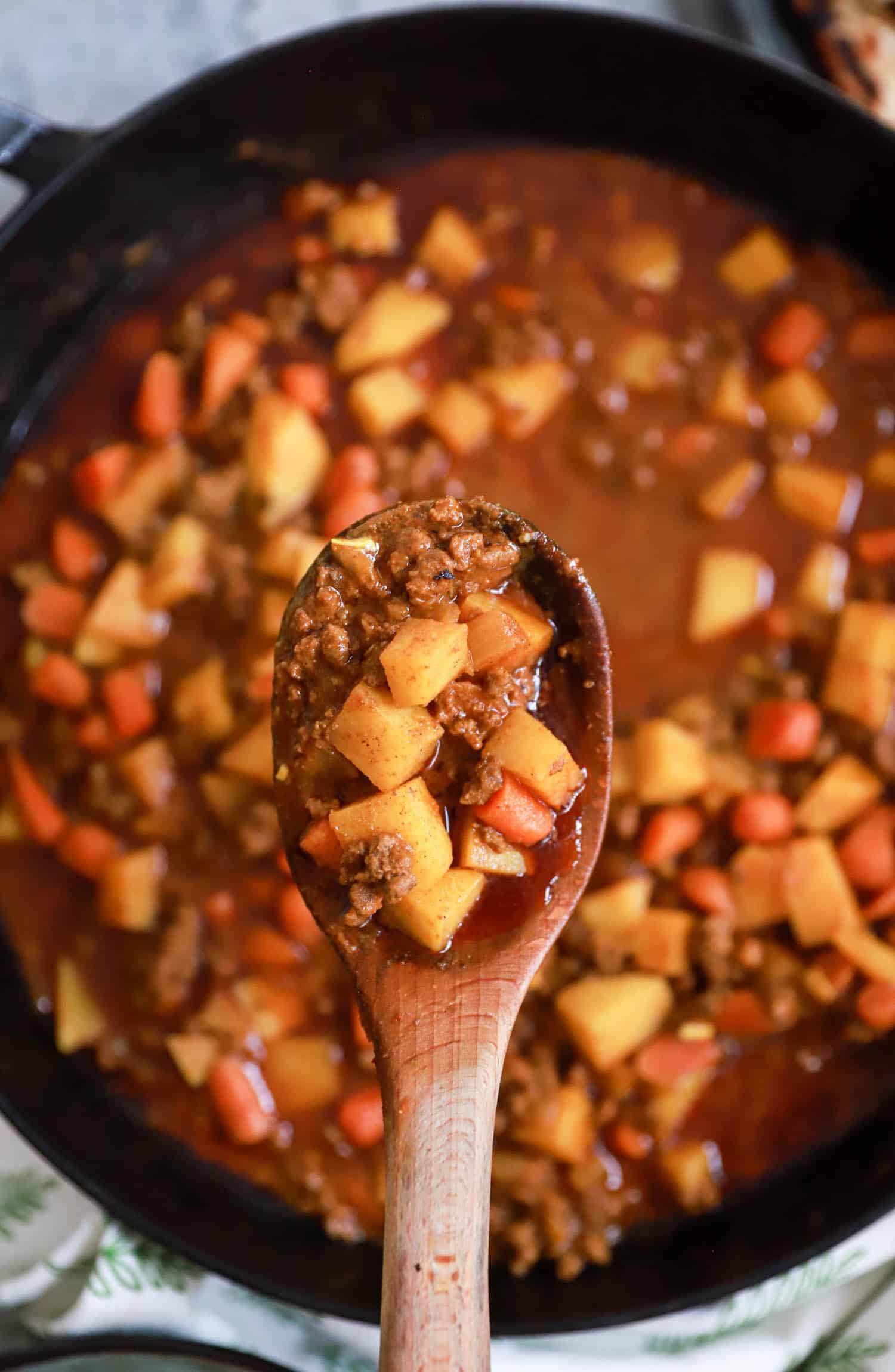 Wooden spoon of beef and potato curry lifted above skillet.