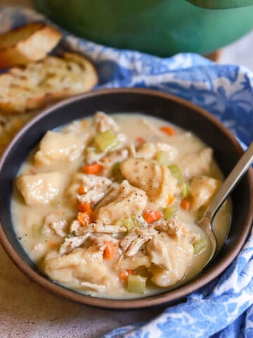 Black ceramic bowl of turkey and dumpling soup with blue dish towel and pieces of baguette.